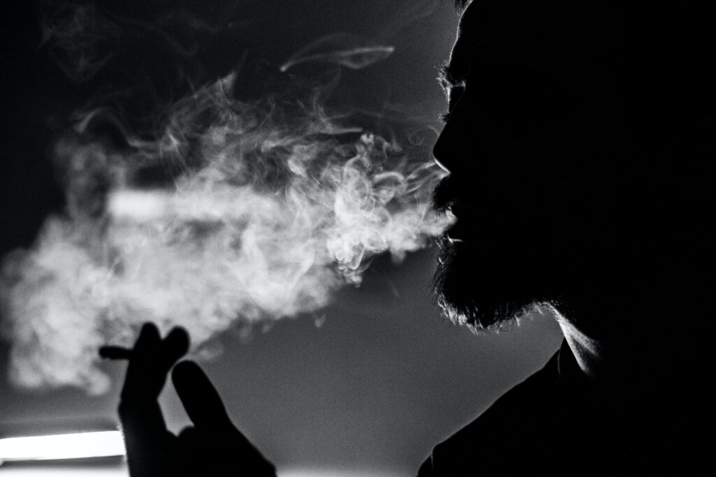 Black and white image. Man holding a cigarette and smoke coming out of his mouth